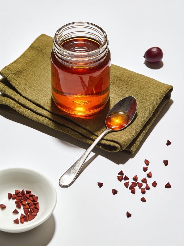 Un frasquito de cristal con aceite de achiote anaranjado rojizo y una cuchara al lado, semillas de achiote enteras regadas sobre la mesa y unas pocas en un platito.
