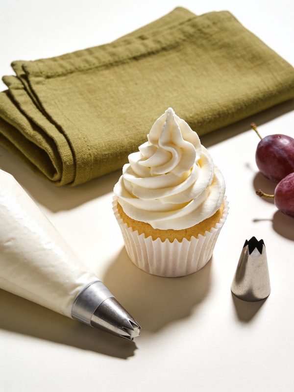 Un remolino de buttercream de vainilla blanco y esponjoso sobre un cupcake de vainilla, con una manga pastelera y una boquilla de estrella al lado.