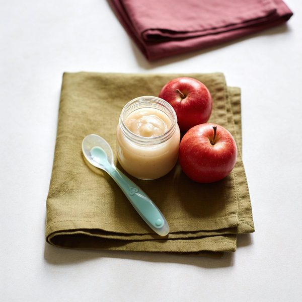 A small glass jar of smooth pale applesauce with a silicone baby spoon and two red apples beside it.