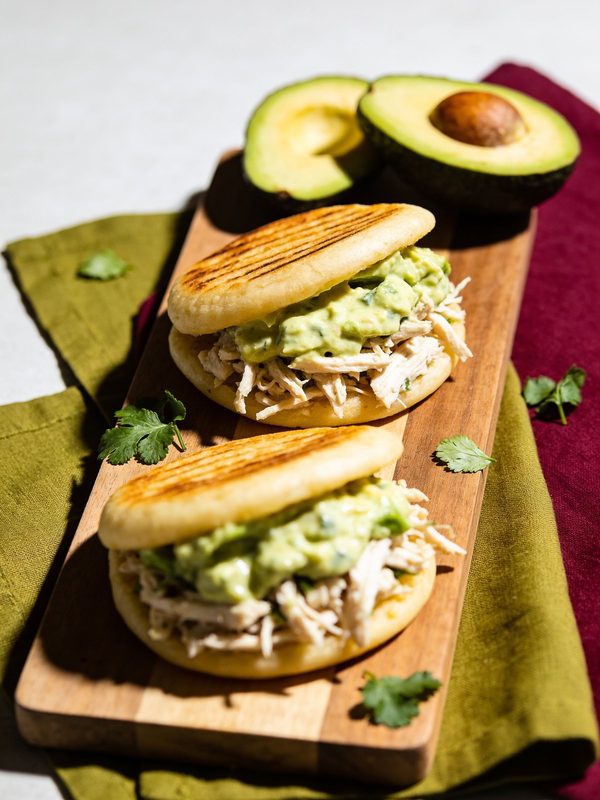 Two grill-marked arepas stuffed with shredded chicken and creamy avocado sauce, with avocado halves and cilantro.