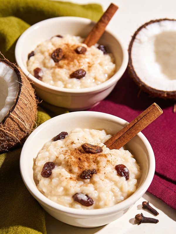 Two bowls of creamy coconut rice pudding with raisins, ground cinnamon and a cinnamon stick, with halved coconuts beside them.