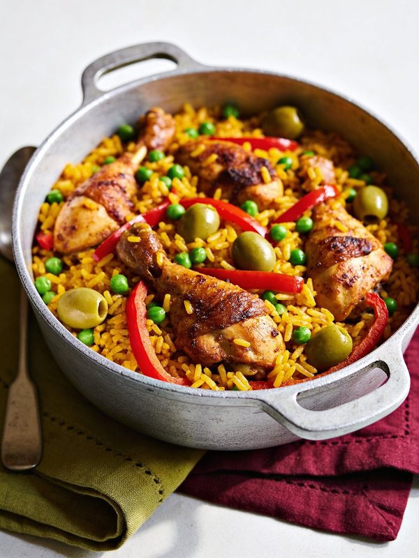 Yellow rice with browned chicken drumsticks, green peas, olives and red pepper strips in a pot.