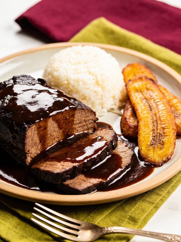 Slices of asado negro in a dark glossy sauce with white rice and fried sweet plantains.