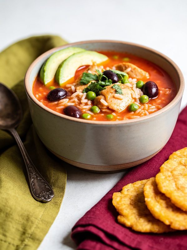 A bowl of soupy rice with chicken, peas and olives in a red broth, topped with avocado and cilantro, with tostones on the side.