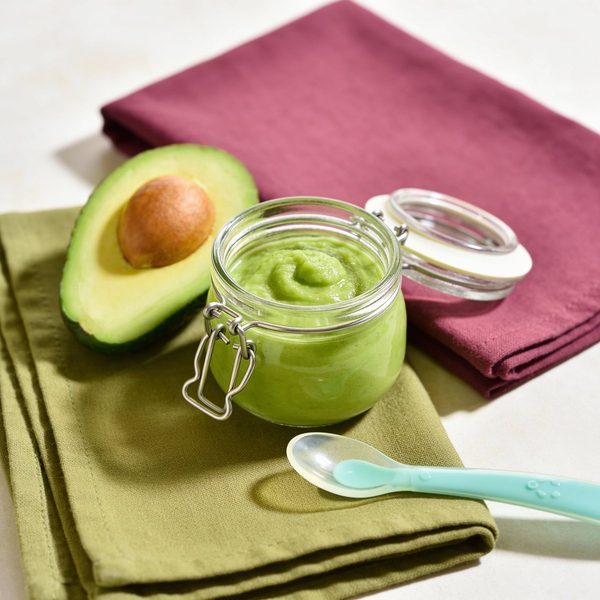 A small glass jar of smooth green avocado mash with a silicone baby spoon and a fresh halved avocado beside it.