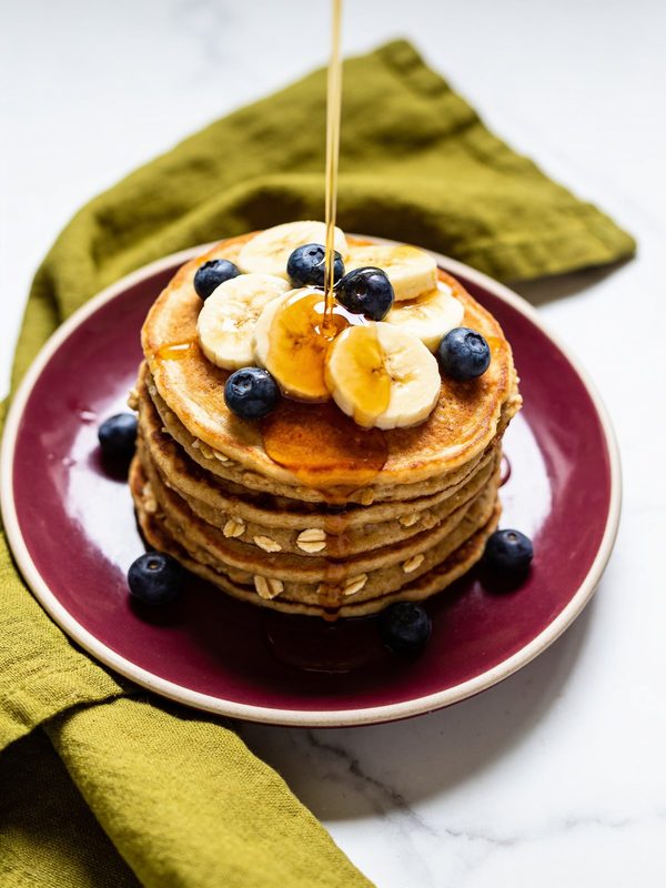 A stack of golden banana oat pancakes topped with banana slices and maple syrup.