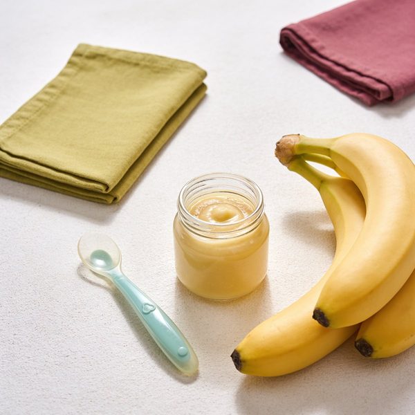 A small glass jar of smooth pale yellow banana puree with a silicone baby spoon and a bunch of fresh bananas beside it.