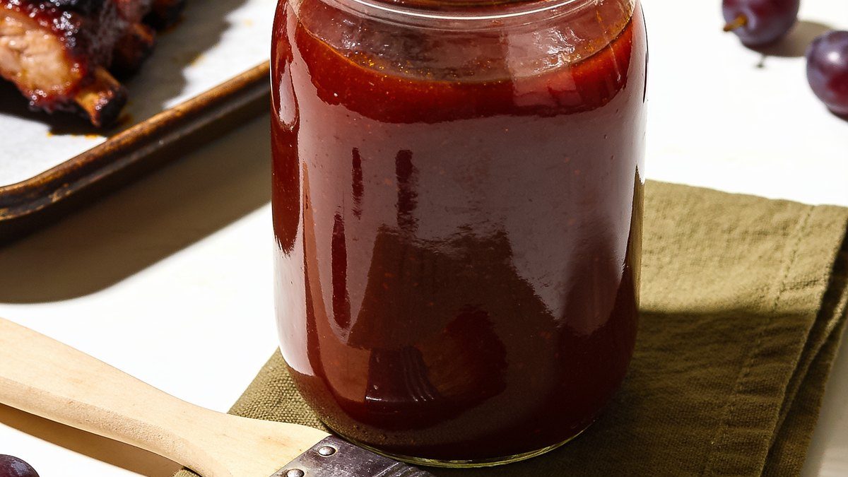 A glass jar of thick, glossy dark red barbecue sauce with a basting brush and grilled ribs beside it.