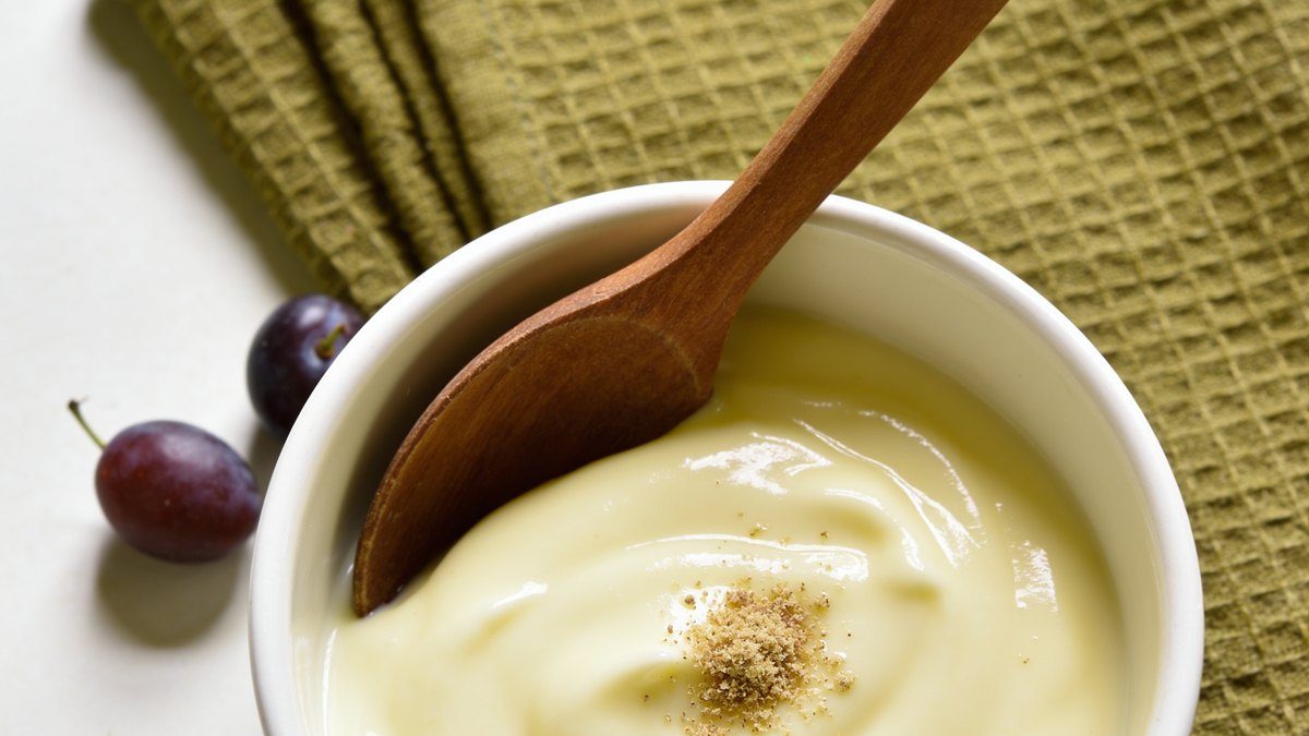 Un tazón blanco con salsa bechamel suave y brillante, una cuchara de madera y un poquito de nuez moscada rallada por encima.