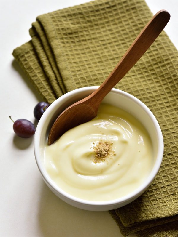 A white bowl of smooth, glossy béchamel sauce with a wooden spoon and a little grated nutmeg on top.
