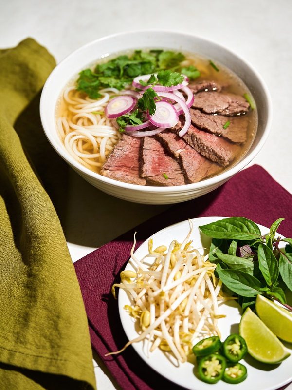 A bowl of beef pho with rice noodles, slices of beef, red onion and cilantro, next to a plate of bean sprouts, Thai basil, lime and jalapeño.
