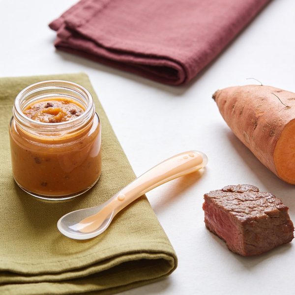 A small glass jar of orange beef and sweet potato puree with a baby spoon, a halved sweet potato and a piece of cooked beef beside it.