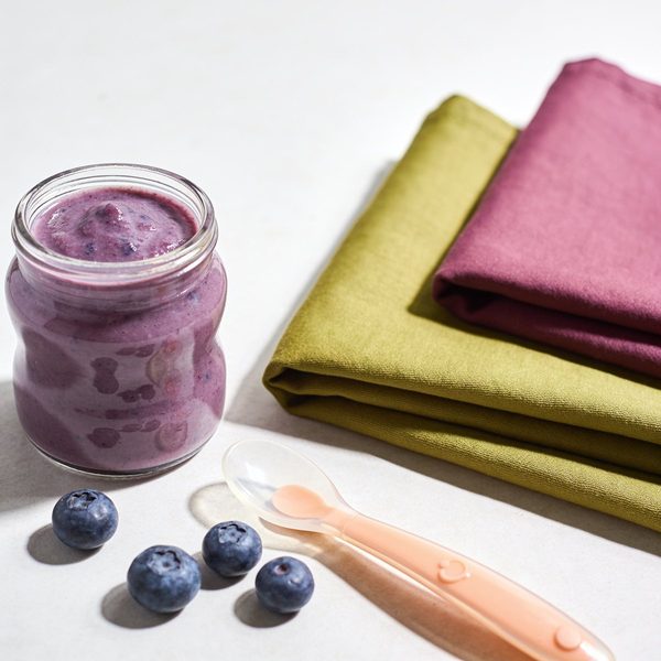 A small glass jar of smooth purple blueberry puree with a silicone baby spoon and a few fresh blueberries beside it.
