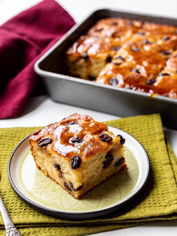 A square of bread pudding with raisins and a glossy caramel top on a plate, with the rest of the pudding in a baking pan behind it.