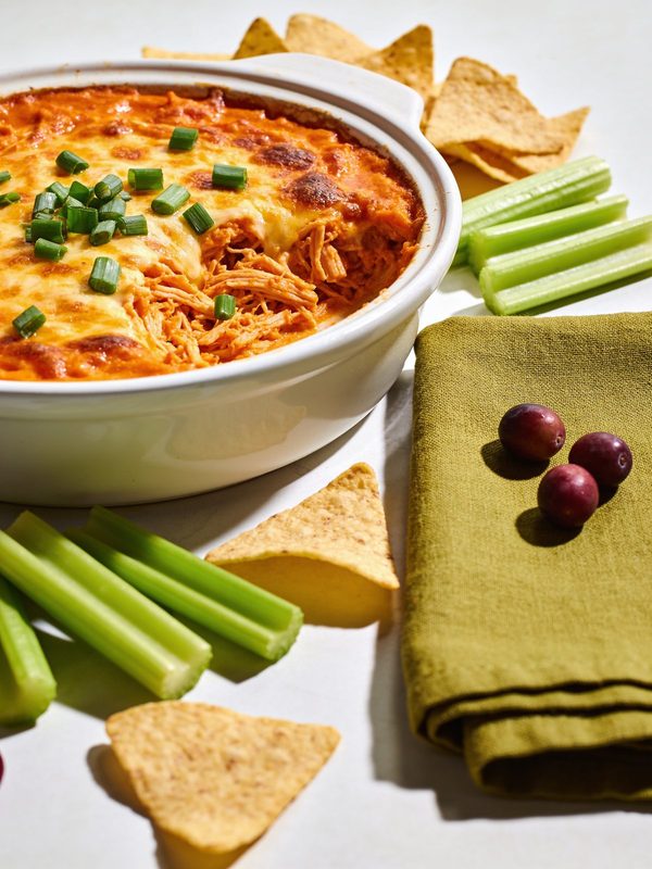 Hot buffalo chicken dip with melted cheddar and green onions in a baking dish, with a chip scooping a cheesy bite, tortilla chips and celery sticks around it.