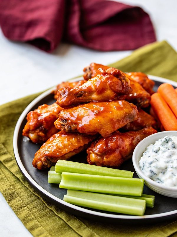 Crispy chicken wings in glossy orange buffalo sauce piled on a dark plate with celery, carrot sticks and a bowl of blue cheese dressing.