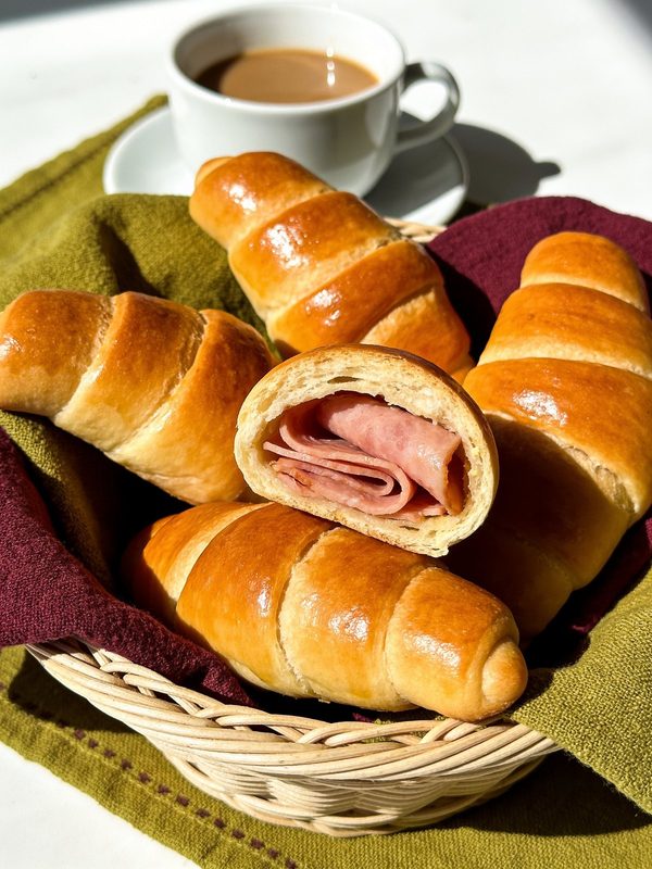A basket of shiny golden cachitos on a green and burgundy cloth, one cut open to show the ham inside, with a cup of coffee behind.