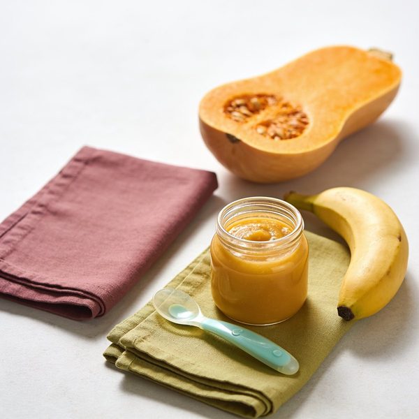 A small glass jar of golden calabaza and plantain puree with a baby spoon, half a squash and a ripe plantain beside it.