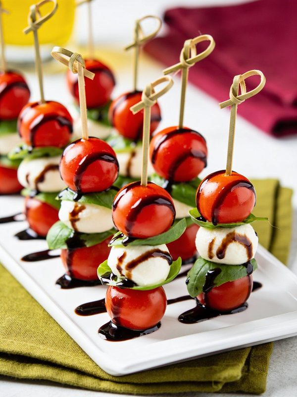 Small skewers of cherry tomatoes, fresh mozzarella and basil drizzled with dark balsamic glaze, lined up on a white platter.