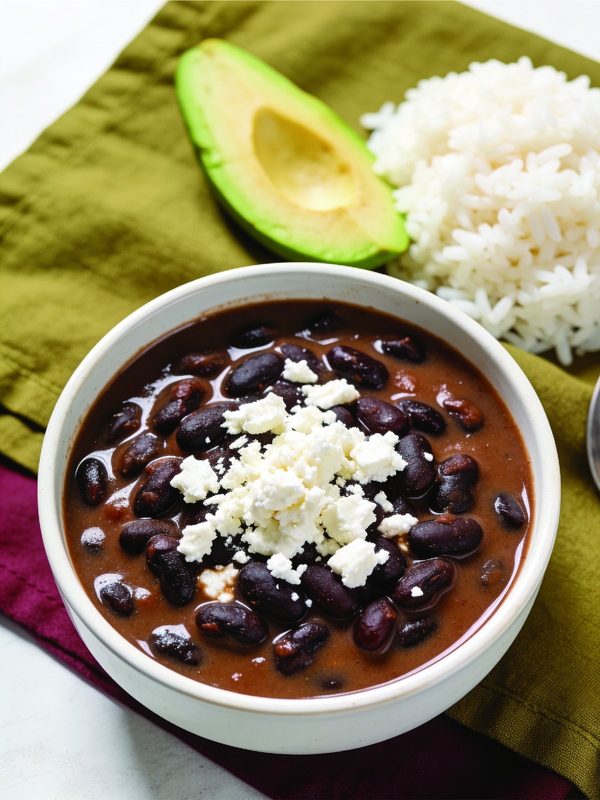 A bowl of Venezuelan black beans topped with crumbled white cheese, with white rice and avocado on the side.