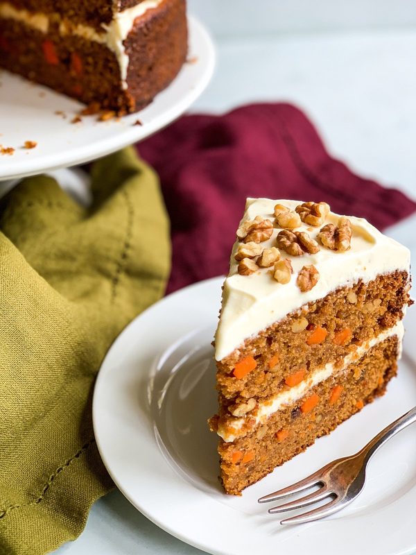A slice of two-layer carrot cake with cream cheese frosting and chopped walnuts on top, with the rest of the cake behind.
