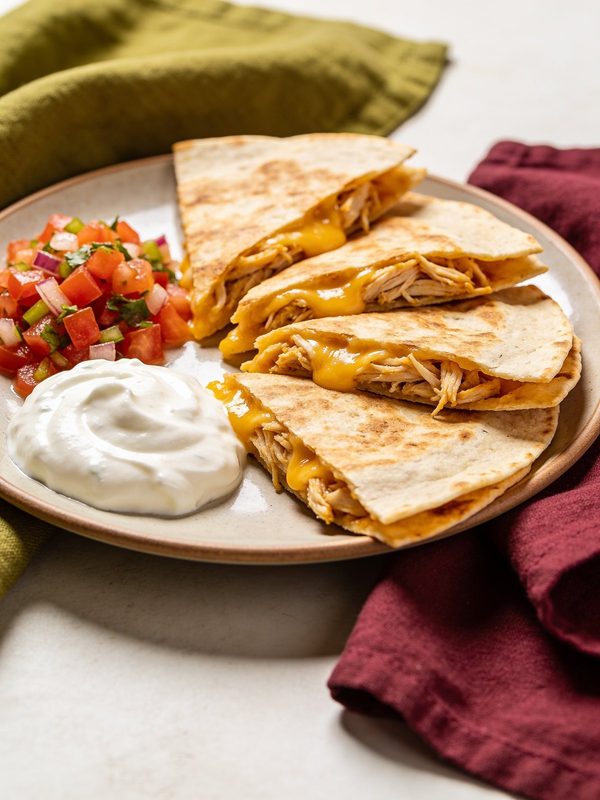 Golden flour tortilla quesadilla wedges filled with shredded chicken and melted cheese, with sour cream and pico de gallo on a plate.