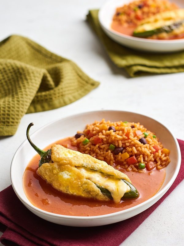 A golden battered chile relleno with melted cheese in a pool of tomato caldillo, served with Mexican red rice with peas, carrots and beans.