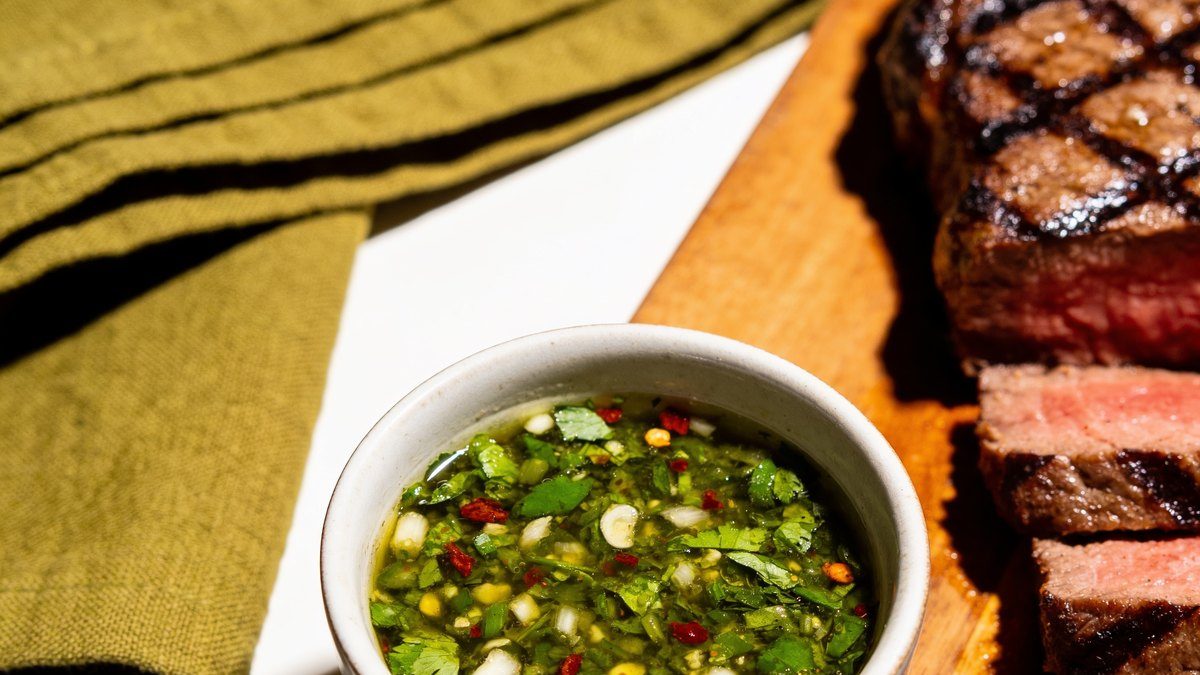 A small bowl of green chimichurri with chopped parsley, garlic and red pepper flakes beside sliced grilled steak.