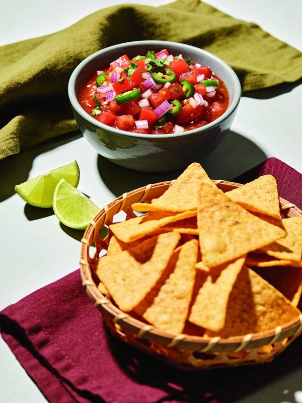 A bowl of chunky red tomato salsa with red onion, jalapeño slices and cilantro next to a basket of golden tortilla chips and lime wedges.