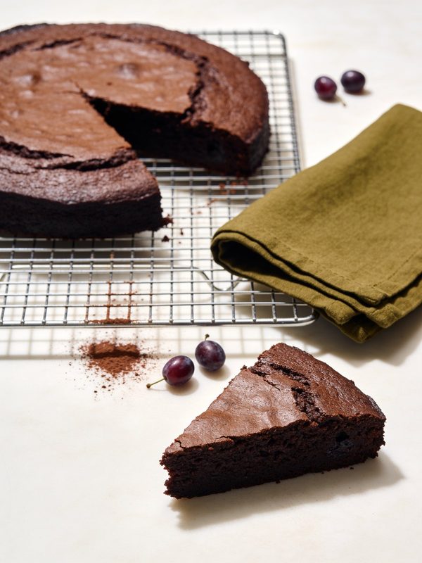 Una capa de bizcocho de chocolate oscuro y húmedo sobre una rejilla, con un pedazo cortado y un poquito de cacao espolvoreado alrededor.