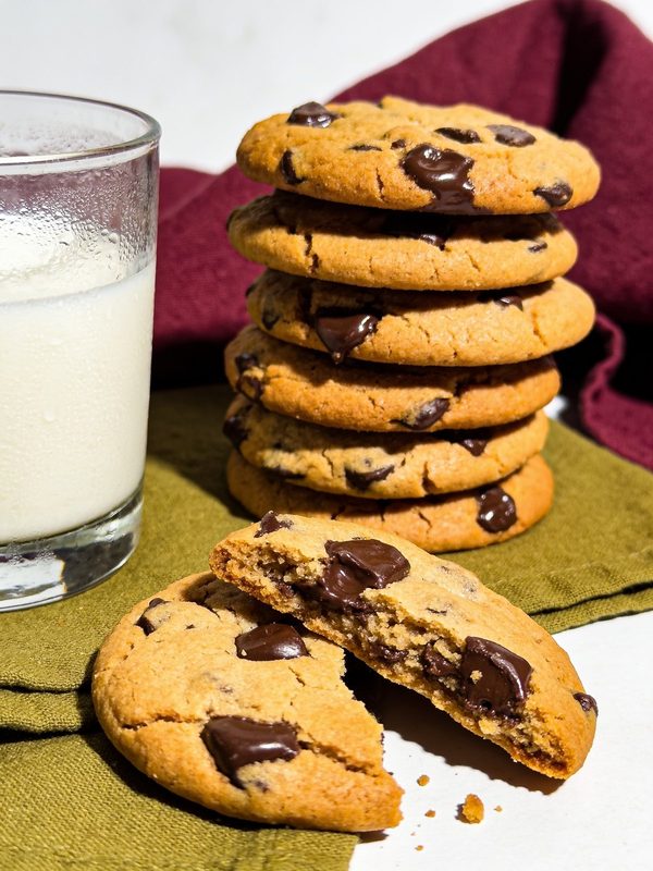 A stack of chocolate chip cookies with melted chocolate chunks, one broken in half, next to a glass of milk.