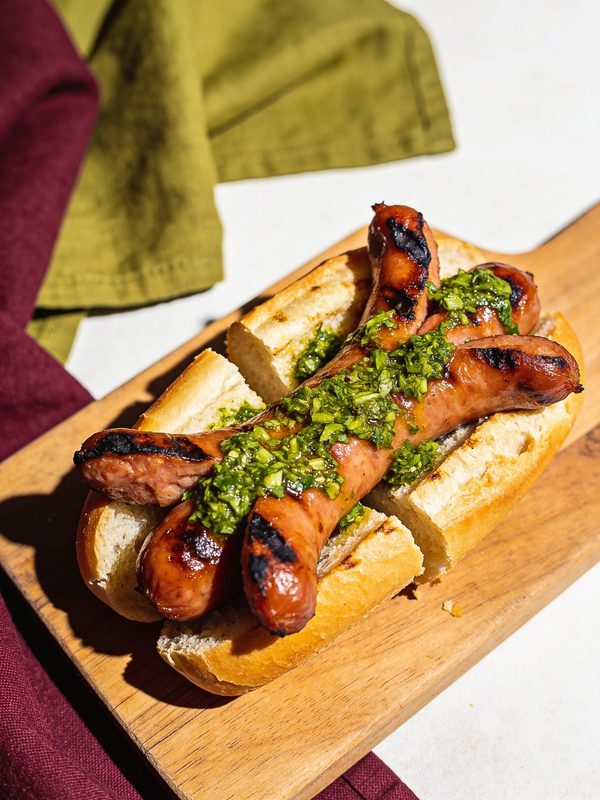 Grilled chorizo sausages with char marks in a split crusty roll, topped with green chimichurri, on a wooden board.