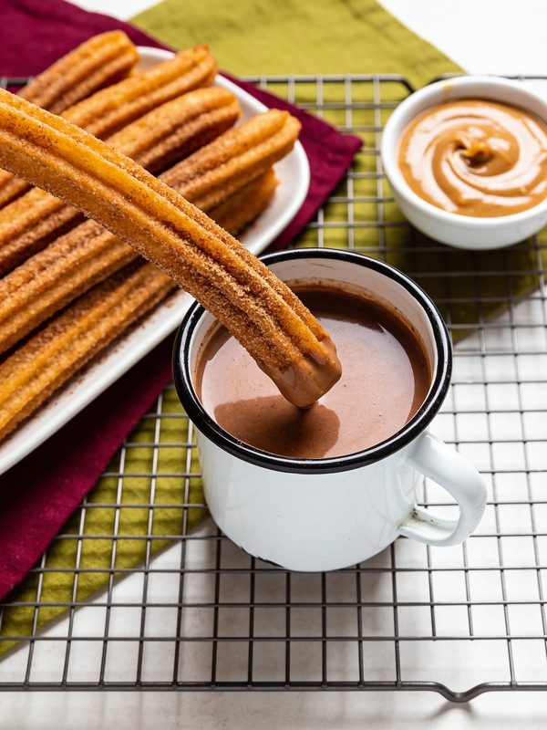 Cinnamon sugar churros on a platter, one dipped in a mug of hot chocolate, with a small bowl of caramel.