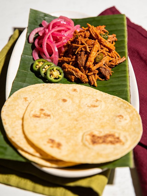 Shredded orange cochinita pibil on a banana leaf with pickled red onions, sliced jalapeño and warm corn tortillas.