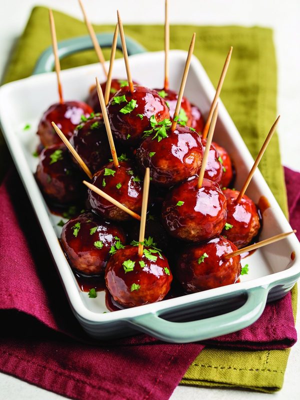 Small meatballs in a glossy dark red sauce, with toothpicks and chopped parsley, in a white serving dish.