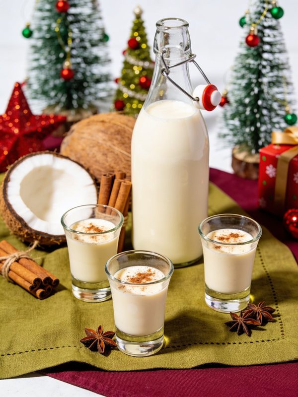 A swing-top bottle of creamy coquito and three small glasses dusted with cinnamon, with coconuts, cinnamon sticks, star anise and Christmas decorations.