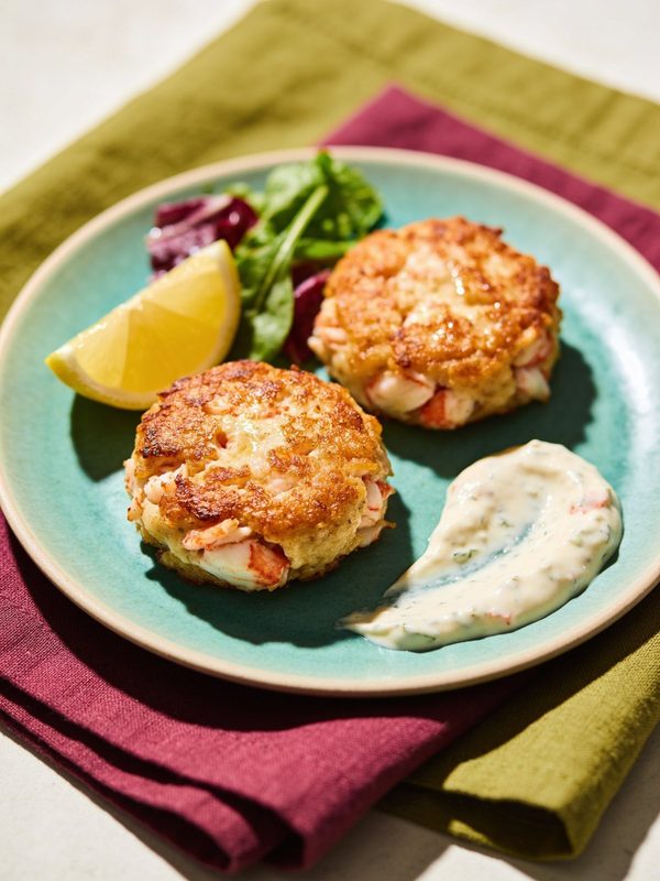 Two golden seared crab cakes with chunks of crab on a teal plate, with a swoosh of remoulade sauce, a lemon wedge and a few mixed greens.