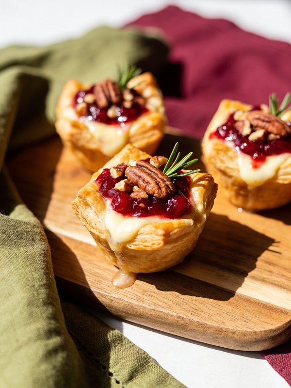 Puff pastry cups with melted brie, cranberry sauce, pecans and rosemary sprigs on a wooden board.