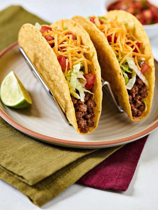 Crunchy corn taco shells standing in a holder, filled with seasoned ground beef, shredded lettuce, diced tomato and shredded cheddar, with a lime wedge and a bowl of salsa.