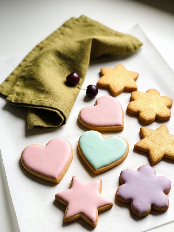Galletas de azúcar en forma de corazón, estrella y flor, algunas decoradas con glaseado en colores pastel, sobre papel de hornear.