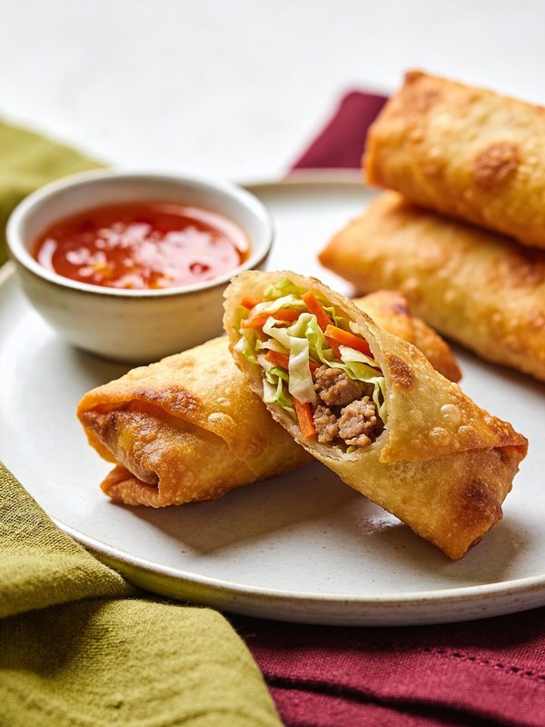 Golden, blistered egg rolls on a plate, one cut open to show shredded cabbage, carrots and ground pork, with a small bowl of sweet and sour sauce.