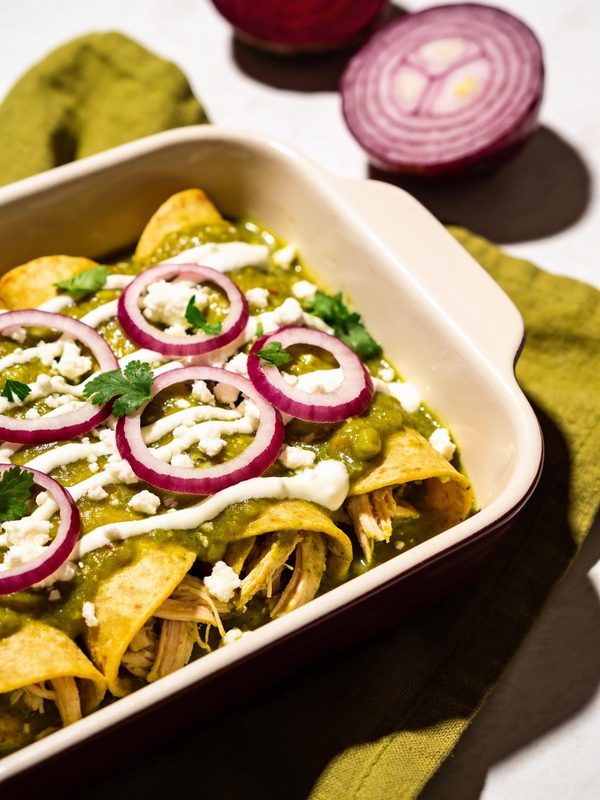 A baking dish of enchiladas verdes filled with shredded chicken, topped with green salsa, crema, crumbled queso fresco, red onion rings and cilantro.