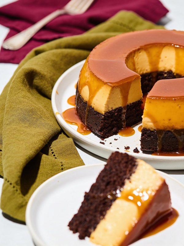 A ring-shaped flancocho with glossy caramel, a creamy flan layer and a dark chocolate cake base, with a slice on a plate in front.
