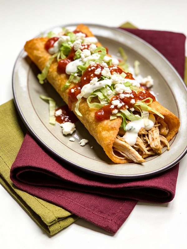 A golden, crisp chicken flauta on a plate, topped with shredded lettuce, crema, crumbled queso fresco and red salsa.
