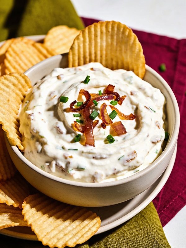A bowl of thick, creamy onion dip topped with caramelized onion strands and chopped chives, surrounded by ridged potato chips.