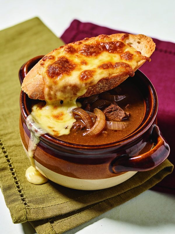 A brown and cream ceramic crock of caramelized onion soup topped with a toasted baguette slice and bubbling, browned melted cheese spilling over the rim.
