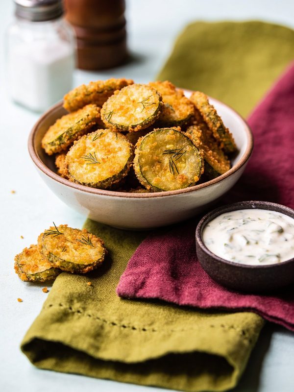 A bowl of crunchy golden fried dill pickle chips garnished with dill, with a small bowl of creamy ranch dressing.