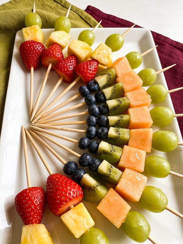 Wooden skewers of strawberries, pineapple, cantaloupe, kiwi, green grapes and blueberries in rainbow order, fanned out on a white platter.