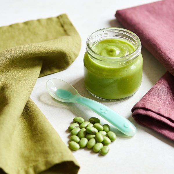 A small glass jar of smooth pale green puree with a silicone baby spoon and fresh green pods beside it.