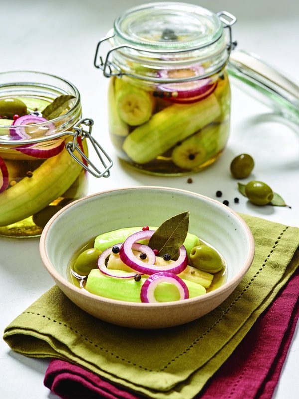 Pickled green banana pieces with red onion rings, green olives, peppercorns and a bay leaf in a bowl, with two glass jars behind it.